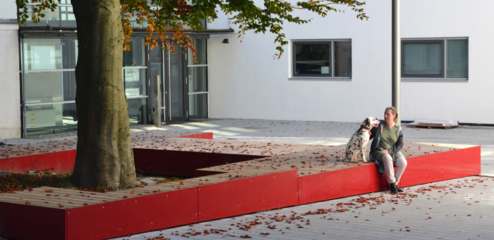 Winkelförmige Sitzbank aus rotem Metall und Holz um einen Baum, eine Person und ein Hund sitzen darauf, von Ney GmbH.