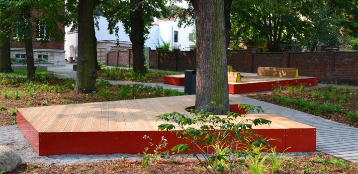 Winkelförmige Sitzbank aus rotem Metall und Holz um einen Baum, ein Design von FreiraumObjekte Ney GmbH.